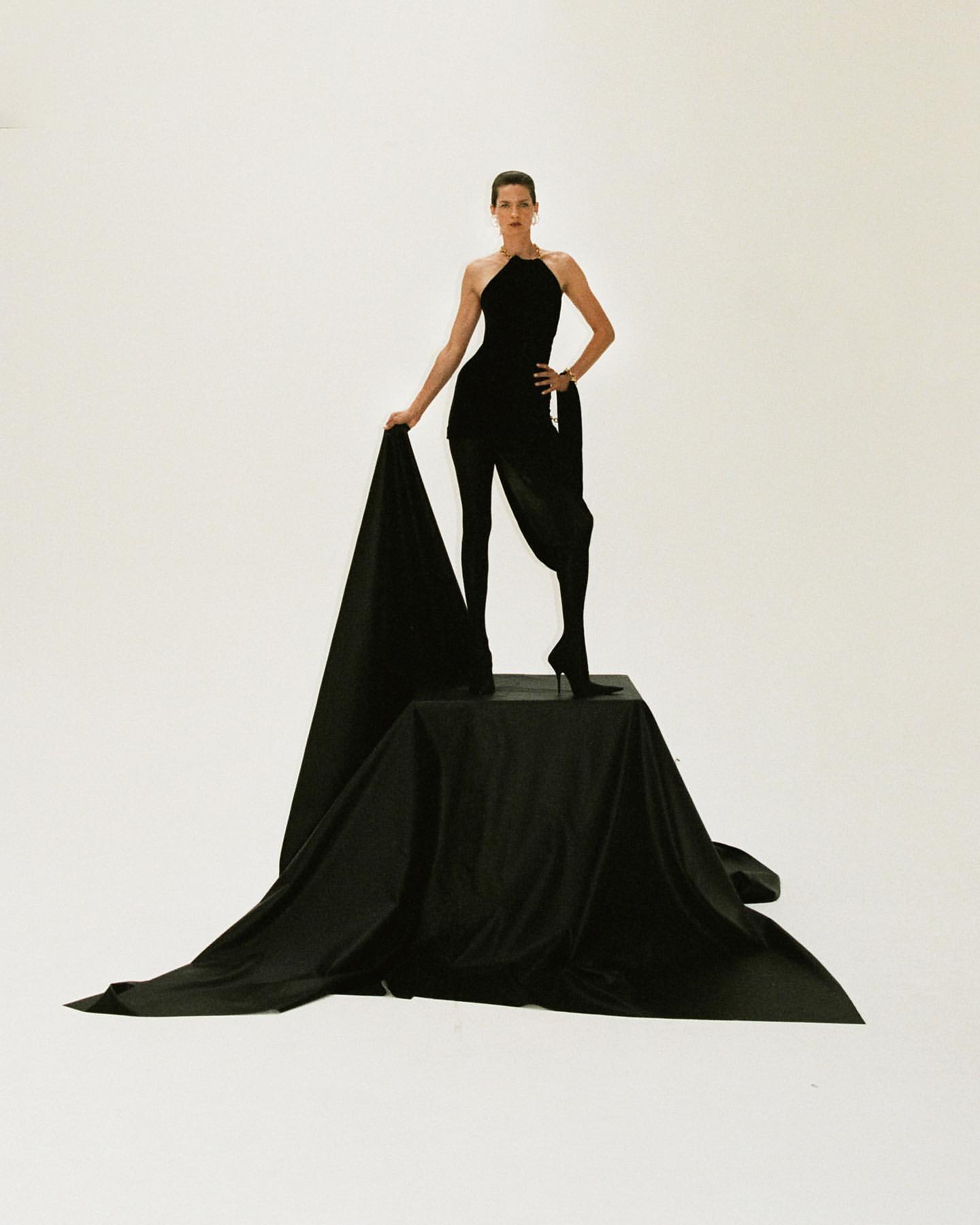 Elegant woman in a black jumpsuit stands on a black platform draped in black fabric against a stark white backdrop in a studio shot.