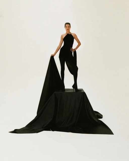 Elegant woman in a black jumpsuit stands on a black platform draped in black fabric against a stark white backdrop in a studio shot.