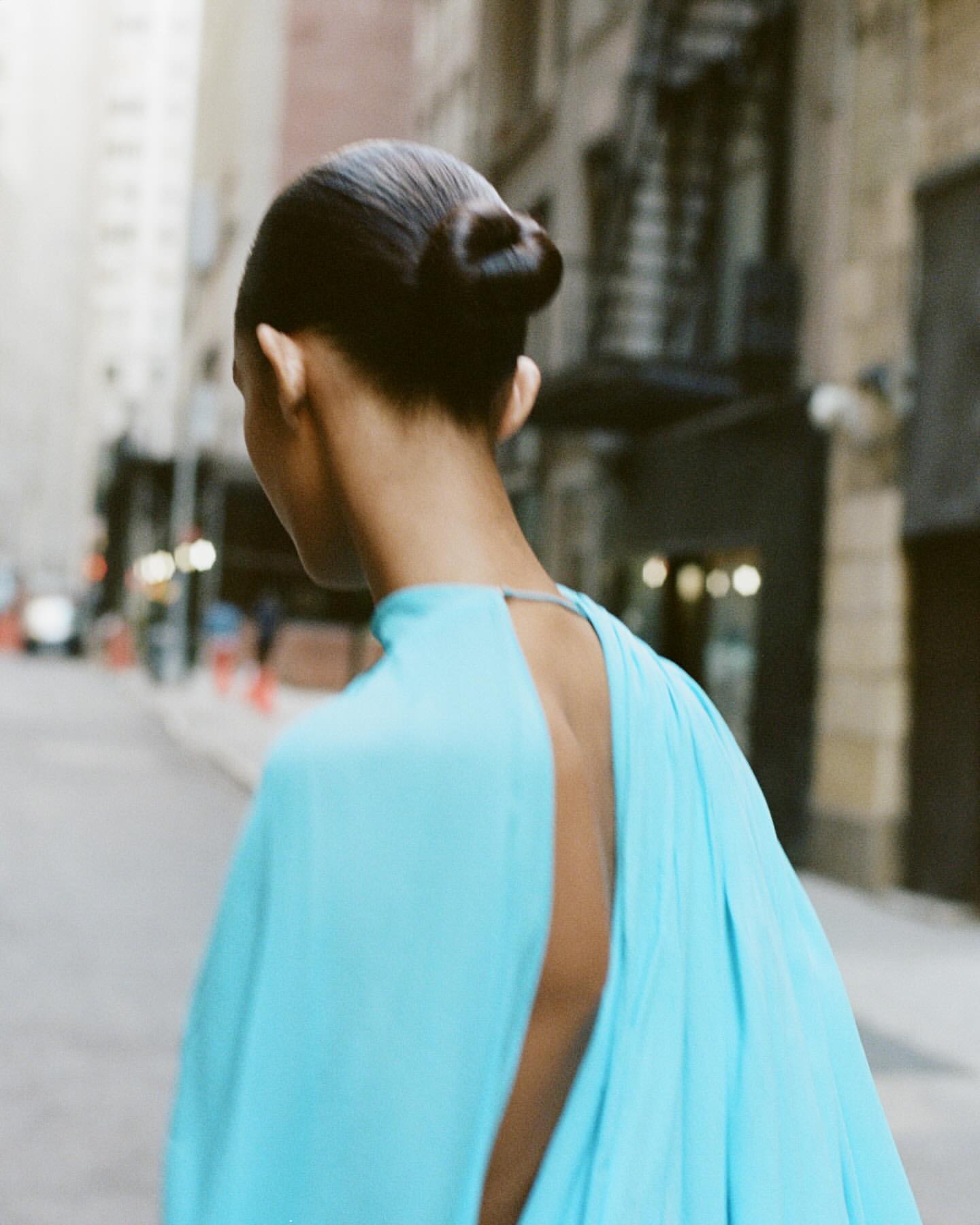 Model with slicked-back hair in a bright blue dress walking down a city street in New York, captured from behind.