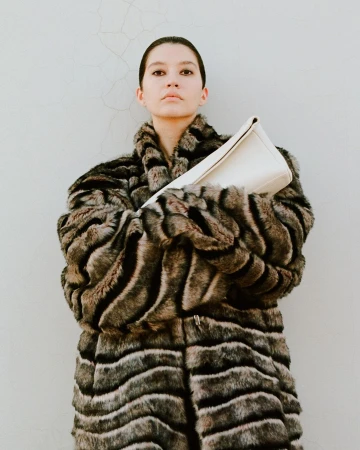 Model wearing a textured brown faux fur coat holding a cream handbag against a pale grey concrete wall.