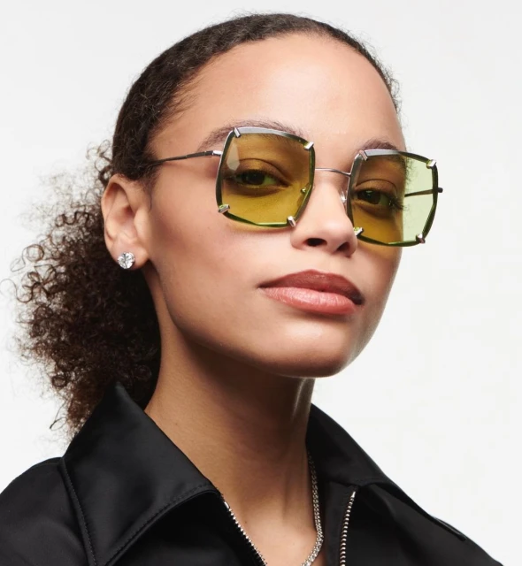 Close-up of a woman with dark curly hair wearing square yellow-tinted glasses, silver chain, and a black jacket against a white backdrop.