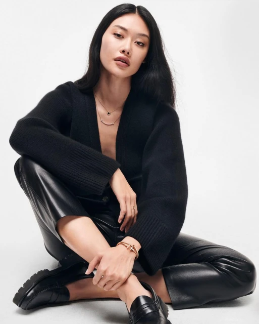 A woman is sitting in black clothing and leather pants in a studio environment. She is wearing jewelry and black shoes.