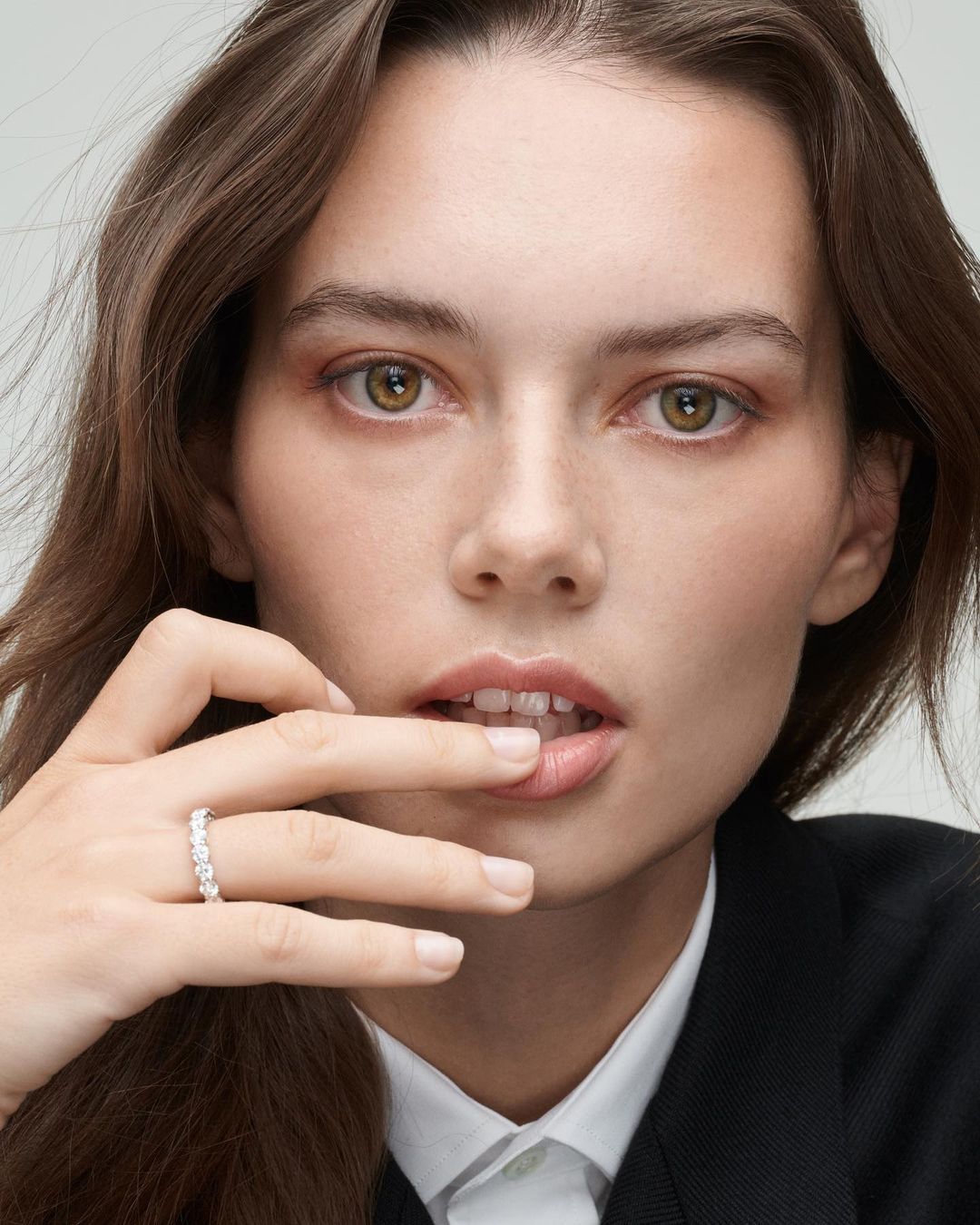 Close-up of a young woman wearing a black blazer and a diamond ring on her finger, with her hand near her mouth.