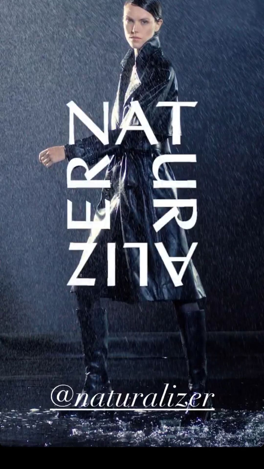 An adult woman poses in a black trench coat and boots, standing in the rain with a dark, wet urban environment in the background.