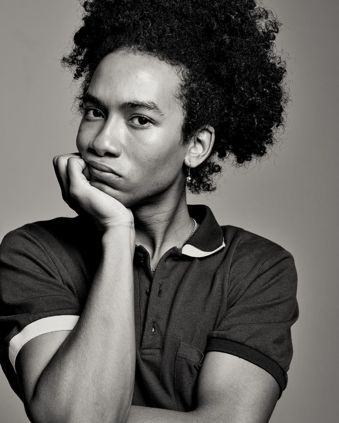 Portrait of a young adult male with an afro resting his chin on his hand, in a studio setting with neutral gray backdrop and diffused lighting.