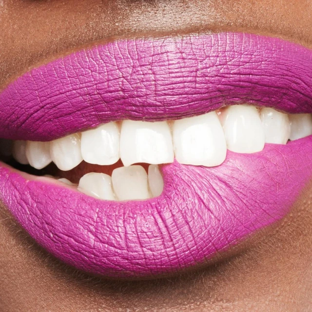 Macro close-up of an adult female with dark skin biting her lip painted with vibrant pink lipstick revealing her white teeth.