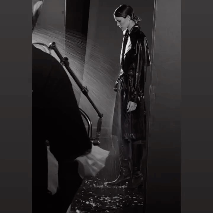 Black and white studio shot of a woman in a dark trench coat, standing in a staged rain environment, and person working the rain machine.