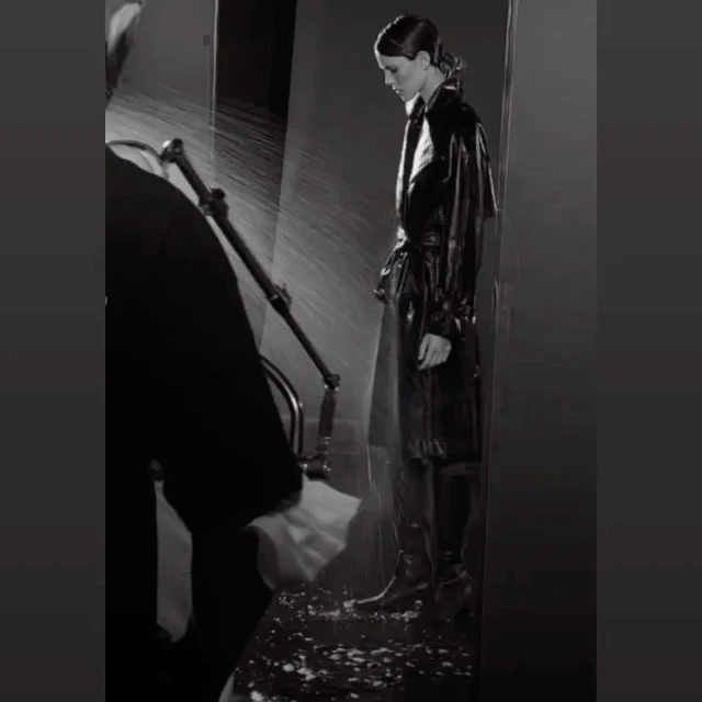 Black and white studio shot of a woman in a dark trench coat, standing in a staged rain environment, and person working the rain machine.