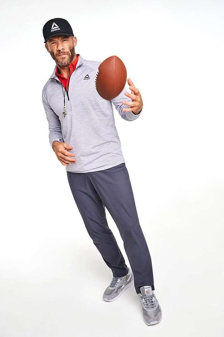 Athletic man in sportswear throws a football in a studio. He wears a black cap and gray athletic wear against a bright white backdrop.
