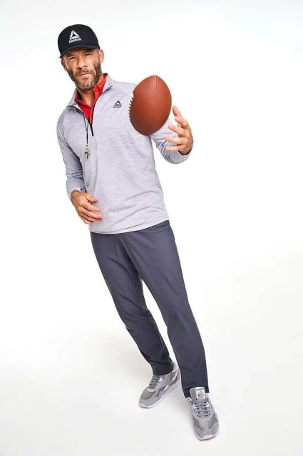 Athletic man in sportswear throws a football in a studio. He wears a black cap and gray athletic wear against a bright white backdrop.