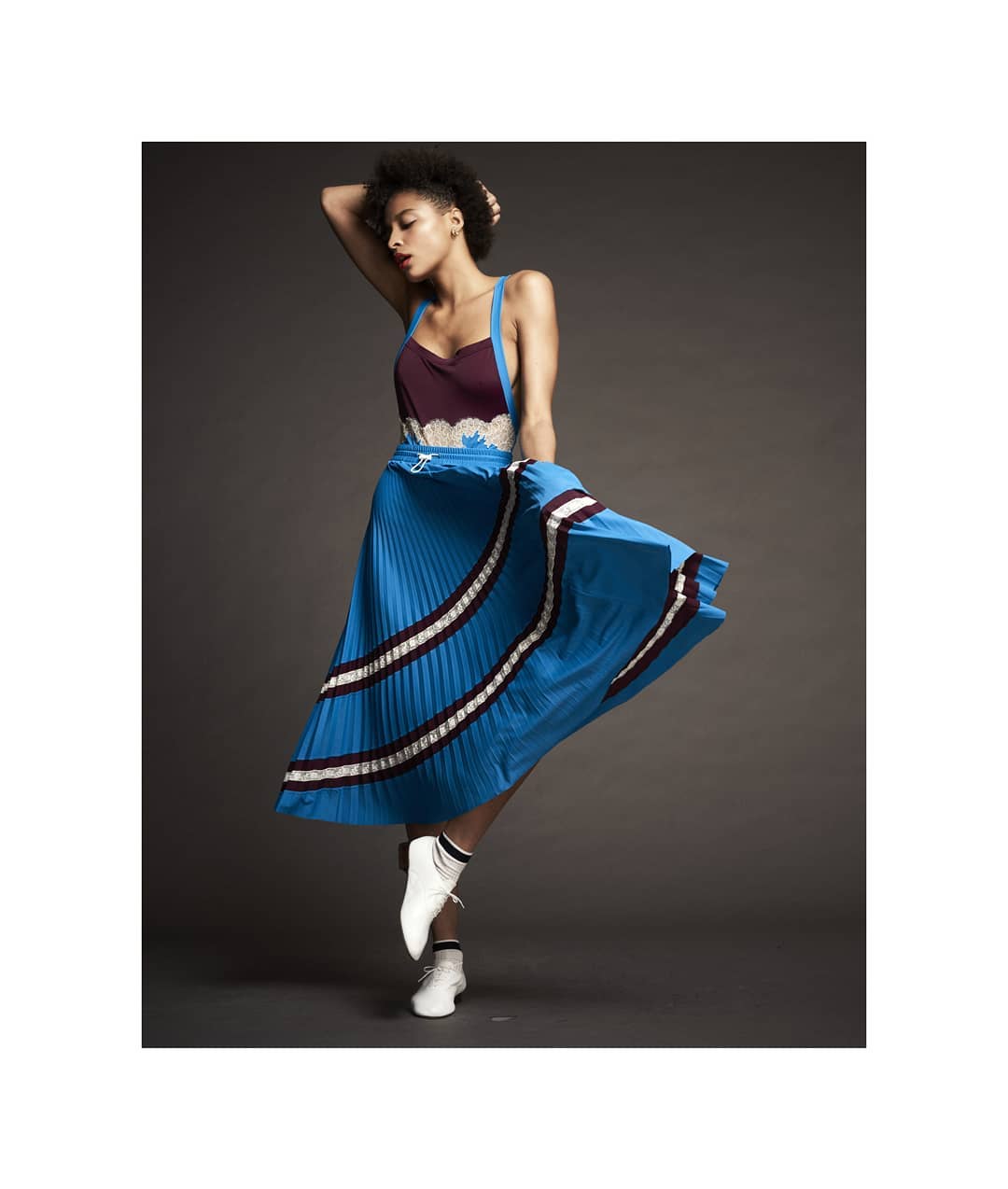 A woman poses in a studio wearing a sleeveless top and a blue pleated skirt with white sneakers against a dark gray backdrop.