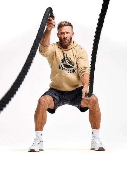 Athletic man in a beige Reebok hoodie works out with black battling ropes in a brightly lit studio setting, looking directly at the camera.