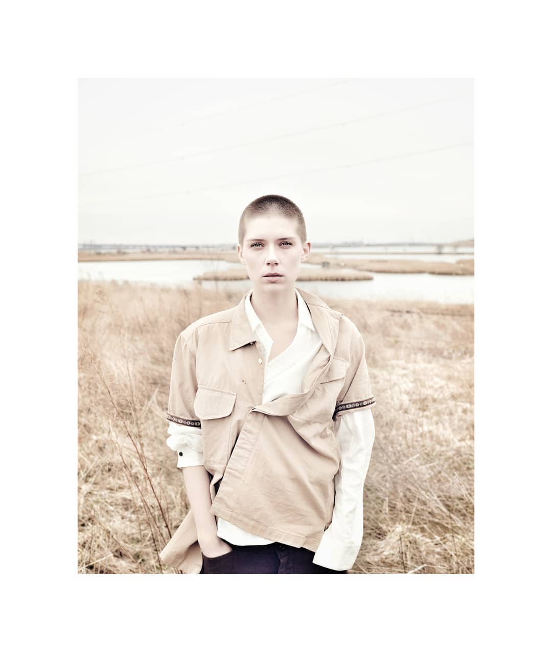 Portrait of a young adult woman with a shaved head posing outdoors in a field wearing a beige shirt and dark jeans in overcast weather.