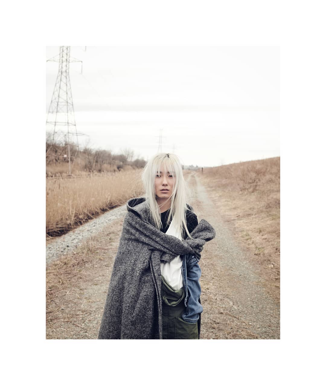 Woman with white hair wearing a hooded cloak stands on a dirt road, evoking a mysterious and ethereal ambiance in a rural landscape.