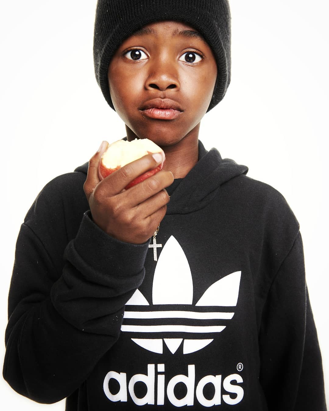 Young Black boy in black beanie and hoodie holding an apple in studio. Close-up portrait with white background, focus on his expression.