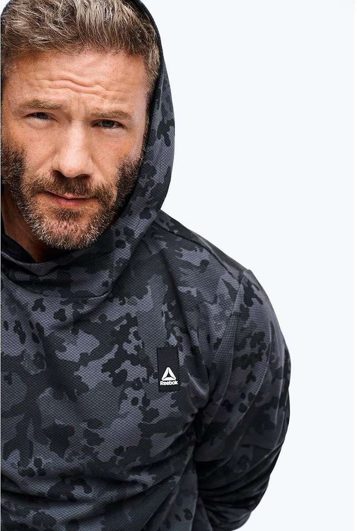 Portrait of a bearded adult male wearing a hooded, gray and black camouflage-patterned sweatshirt, isolated on white background.