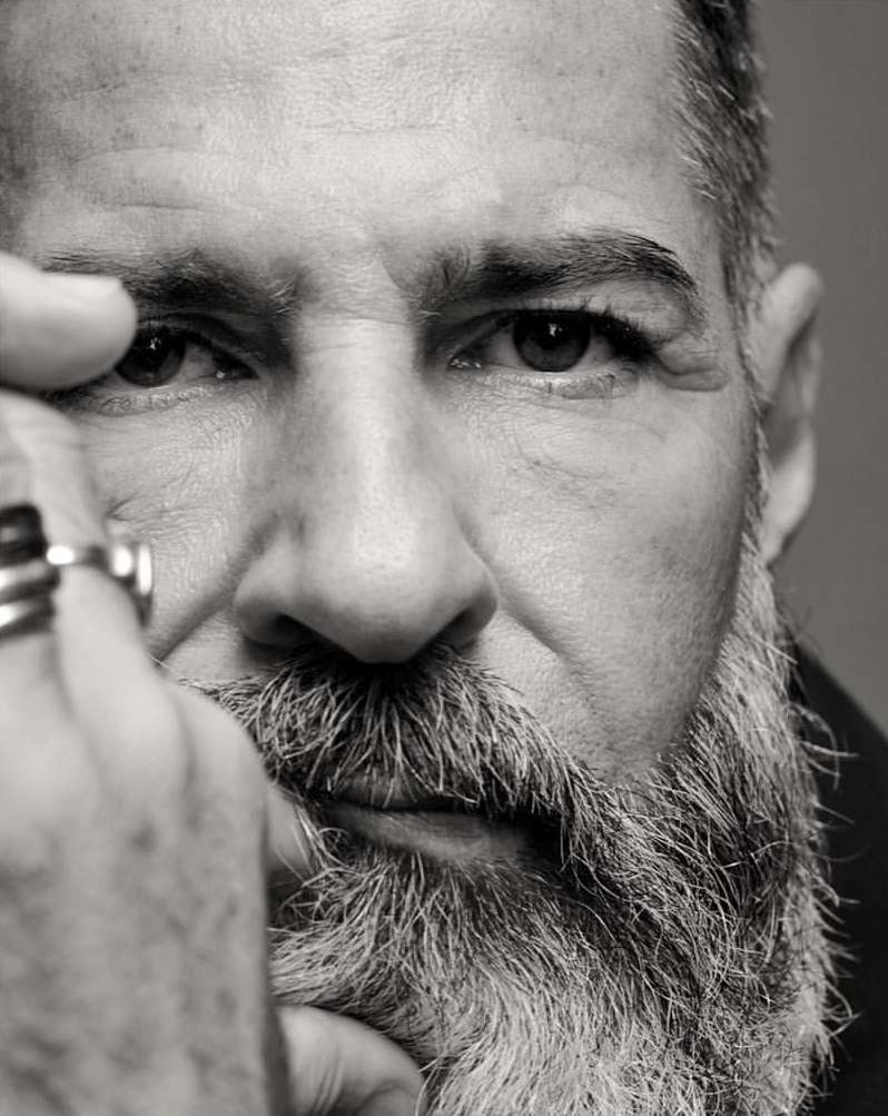 Monochrome portrait of a bearded man with a hand near his face, focusing on the face, eye contact and unique facial hair.