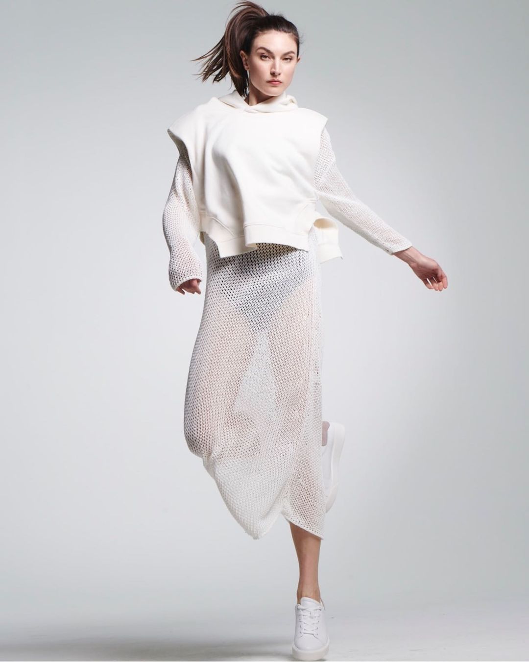 A young adult fashion model poses indoors in a white textured hooded sweatshirt and skirt, jumping in the air with a ponytail.