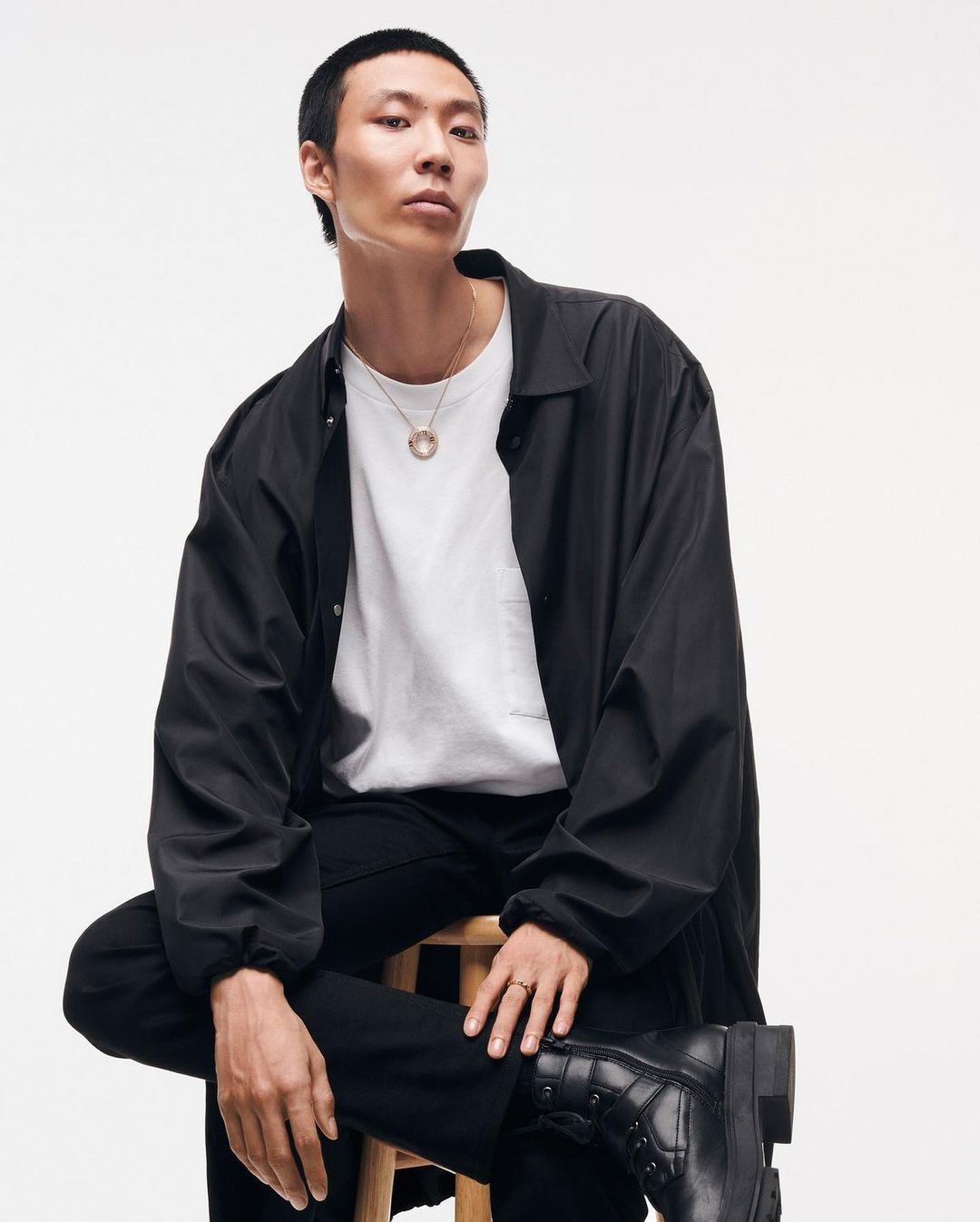 Man wearing a black jacket and white t-shirt sitting on a wooden stool against a white background