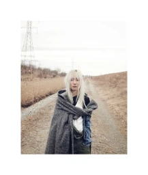 A person with long white-blonde hair wrapped in a grey tweed coat standing on an outdoor dirt path beneath an overcast sky.