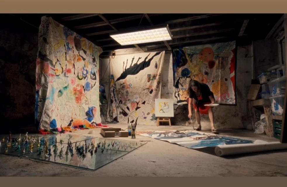 Female artist in studio, painting on large canvas spread on floor. Artwork hangs as backdrop in dimly lit, raw space.