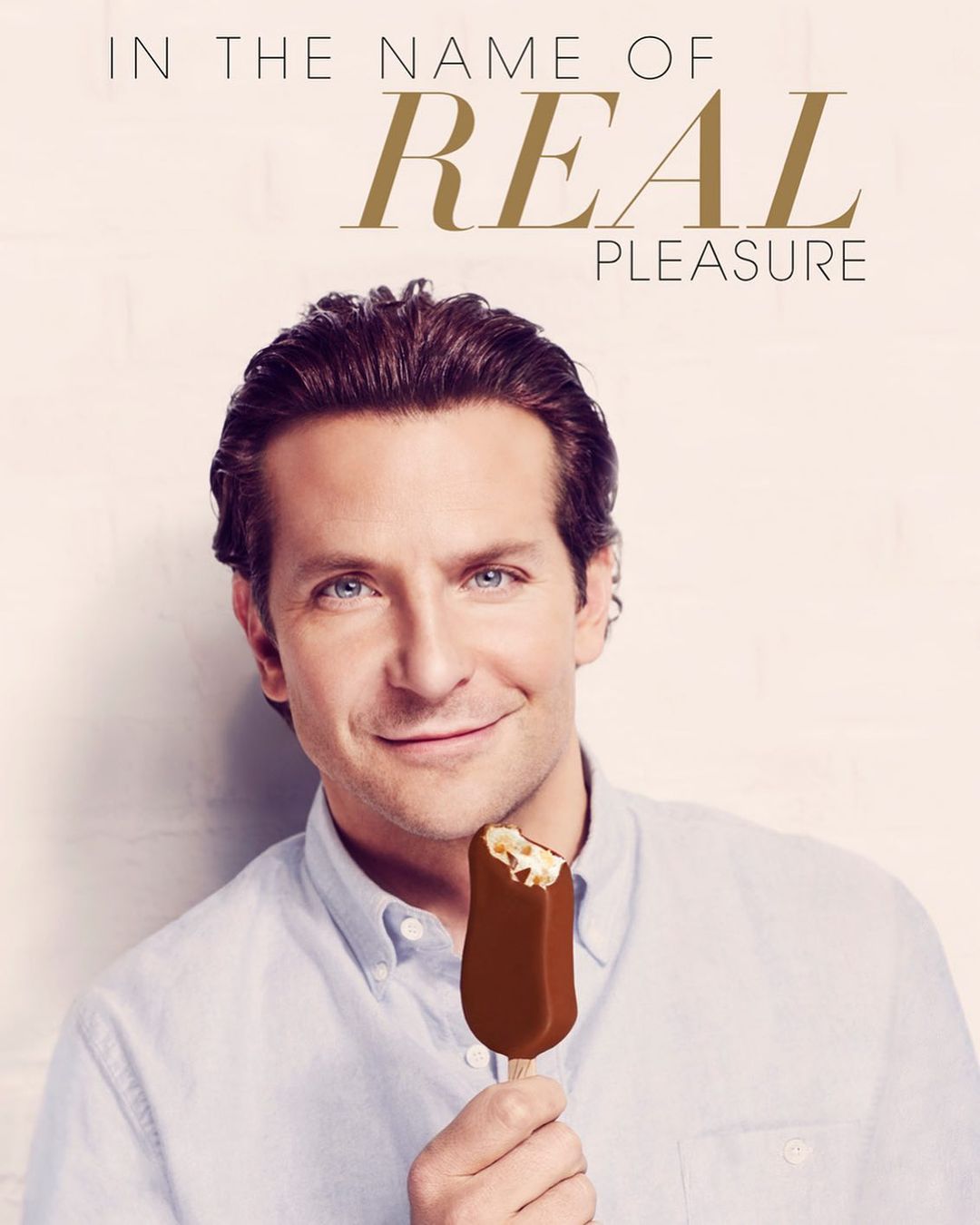 Portrait of Bradley Cooper holding a bitten chocolate ice cream bar against a soft neutral backdrop with advertising text.