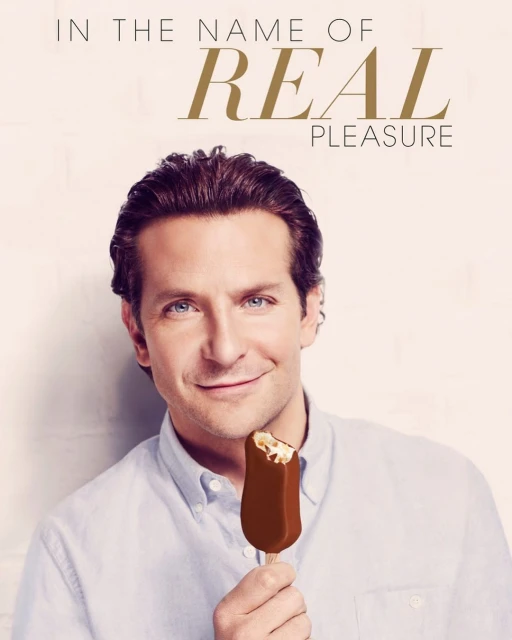 Portrait of Bradley Cooper holding a bitten chocolate ice cream bar against a soft neutral backdrop with advertising text.