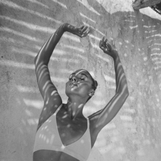 Woman in a bikini top poses in dappled sunlight against a light-colored wall, arms raised, in a monochromatic outdoor shot with soft lighting.