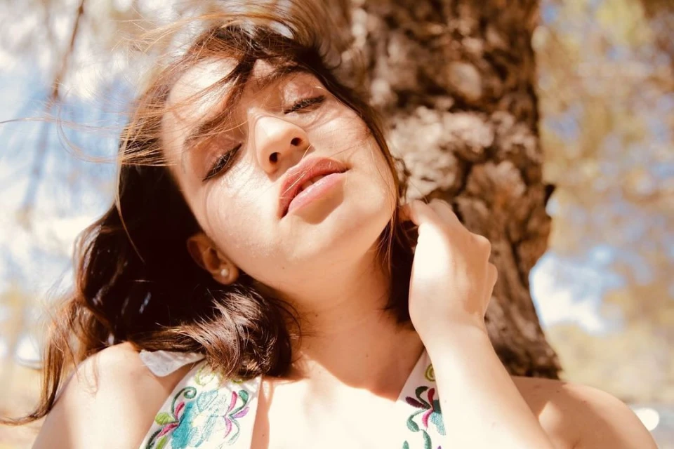 Close-up of a fair-skinned adult female with closed eyes and dark brown hair, outside in natural light with foliage in the background.
