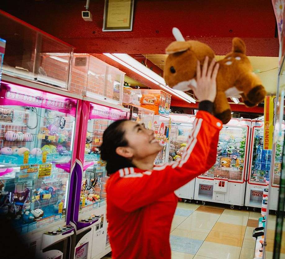 Smiling woman in red track jacket holds up a tan plush deer inside an arcade filled with claw machine games, creating a joyful scene.