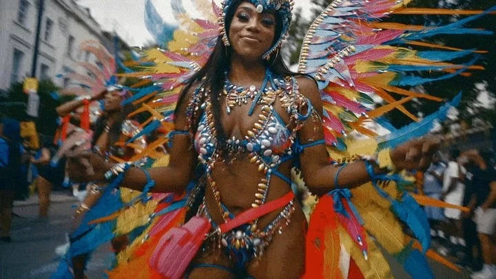 Woman in a colorful, jeweled costume with feathered wings parades in a street, part of a group during a daytime outdoor event.