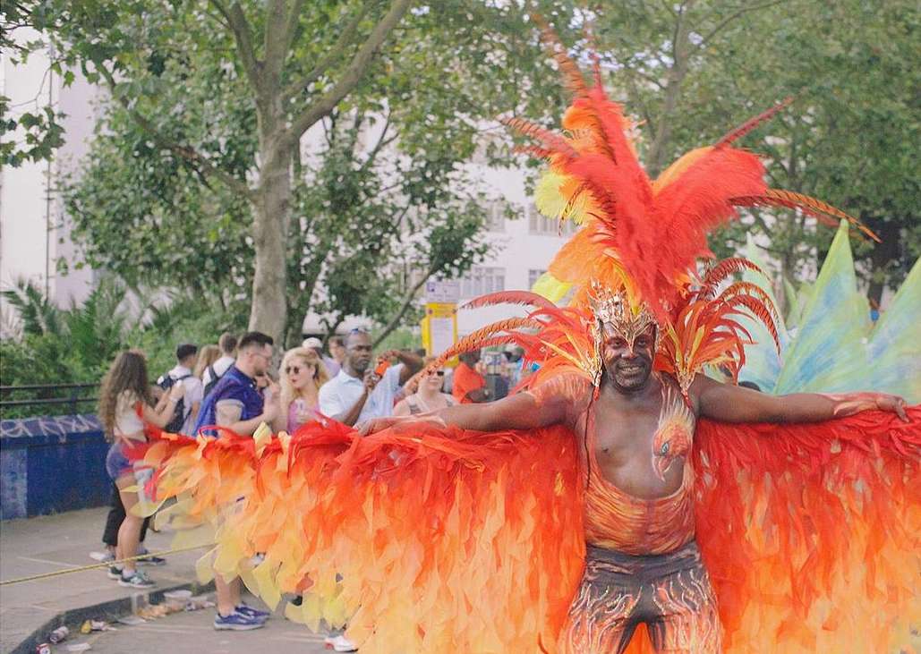 Man with vibrant, colorful feathered costume smiles outdoors amongst a crowd during a lively carnival parade on a sunny day in the city.
