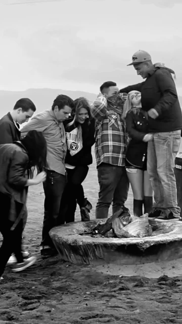 Black and white shot of a group of seven young adults gathered around a stone fire pit on a beach, with mountains in the background.