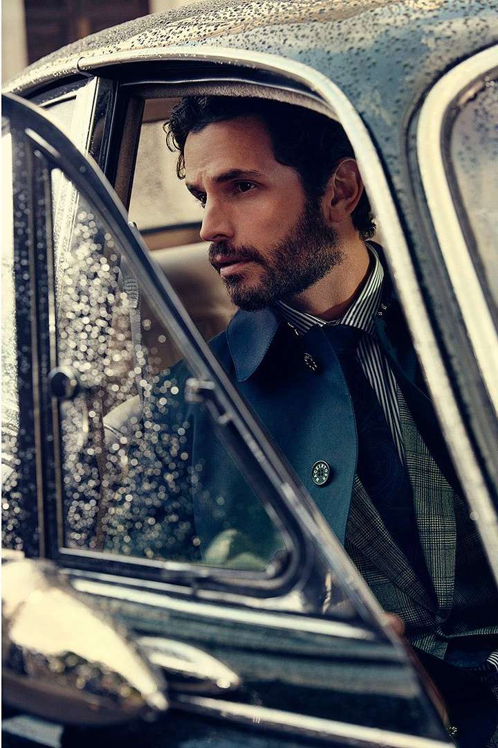 A man exits a vintage car with a serious expression, wearing a navy coat, striped shirt, and dark tie, captured in natural light.