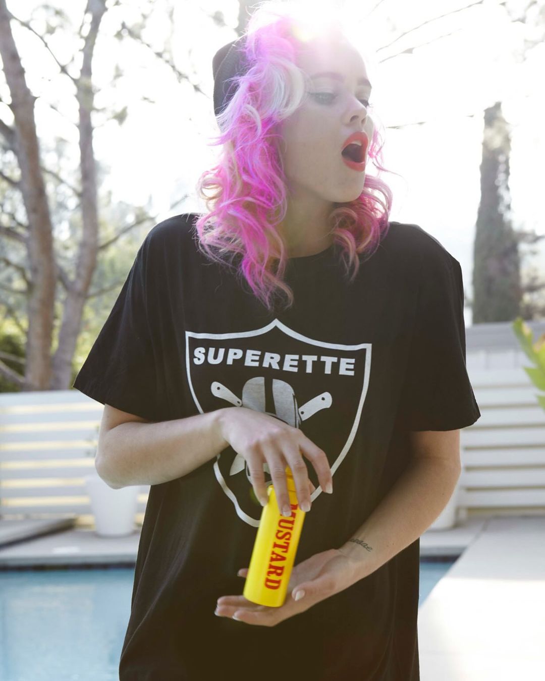 Woman with pink hair wearing a black t-shirt with white logo, holding a mustard bottle in front of a pool in outdoor setting.