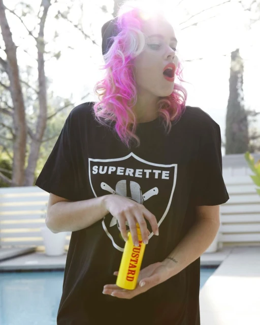 Woman with pink hair wearing a black t-shirt with white logo, holding a mustard bottle in front of a pool in outdoor setting.