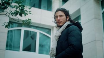 Man with long dark curly hair wearing a jacket and scarf stands outdoors in front of an off-white building with green tree branches visible.