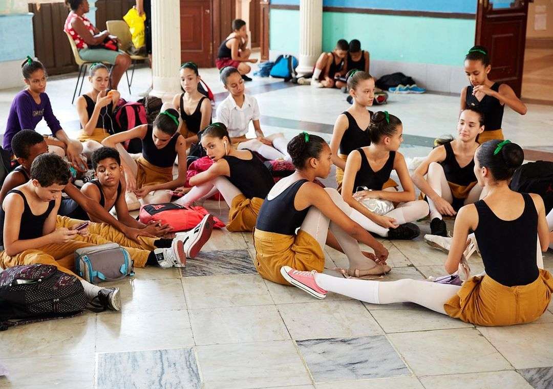 A large group of young ballet students sits on the floor of a dance studio, wearing ballet attire and relaxing between dance routines.