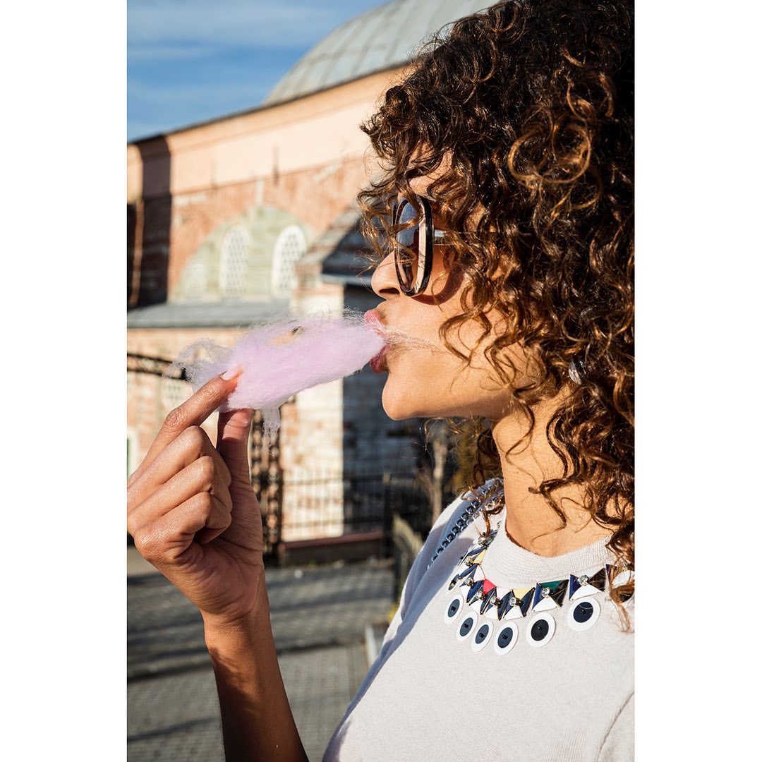 Young woman with curly hair eats pink cotton candy in front of an ornate brick building. She wears sunglasses and a geometric necklace.