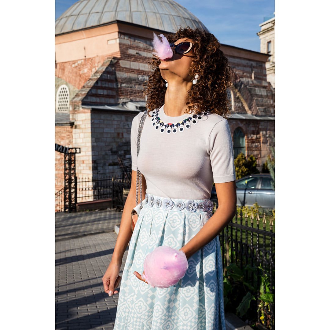 Woman outdoors wearing sunglasses eating pink cotton candy in front of a domed building and parked car on a sunny day.