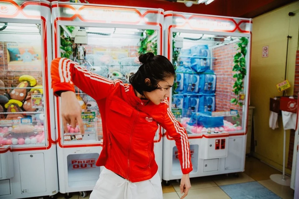 Young adult in red Adidas jacket poses in front of claw crane arcade games with colorful plush toys and blue wrapped water bottles.