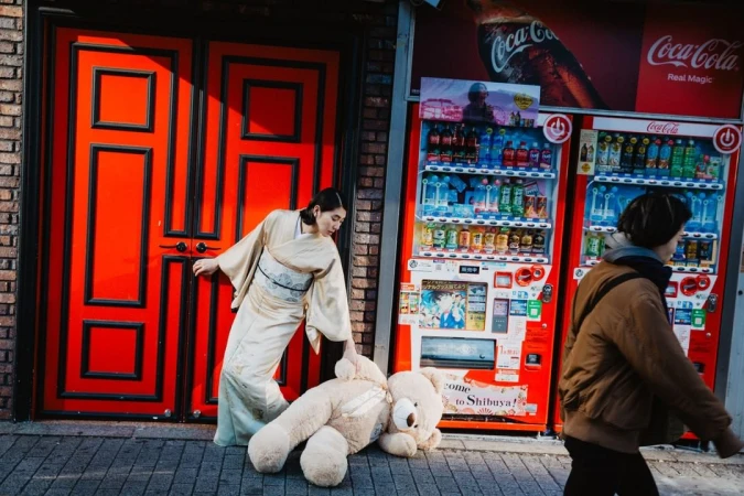 Woman in kimono with teddy bear next to vending machines and red door in urban setting with warm sunlight. Fashion editorial.