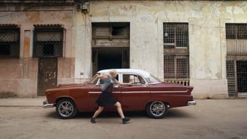 Young woman in a black dress walks in front of a vintage car on a street in Havana, Cuba, with old buildings in the background.