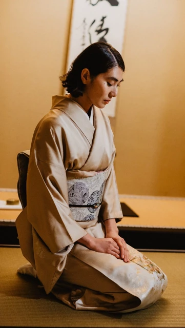 Woman in kimono kneeling indoors with calligraphy scroll in the background in a serene Japanese-style setting, emphasizing Japanese culture.