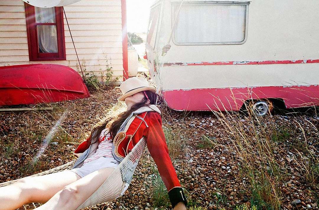 Woman in vintage red jacket resting in a hammock near a white caravan and house, sunlit, casual outdoor scene.