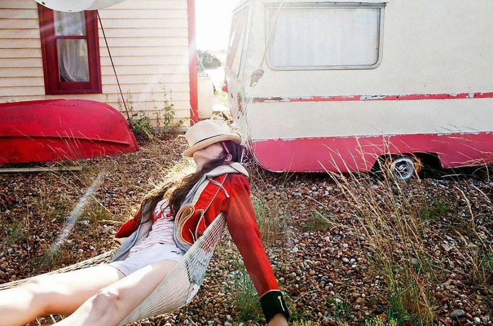 Woman in vintage red jacket resting in a hammock near a white caravan and house, sunlit, casual outdoor scene.