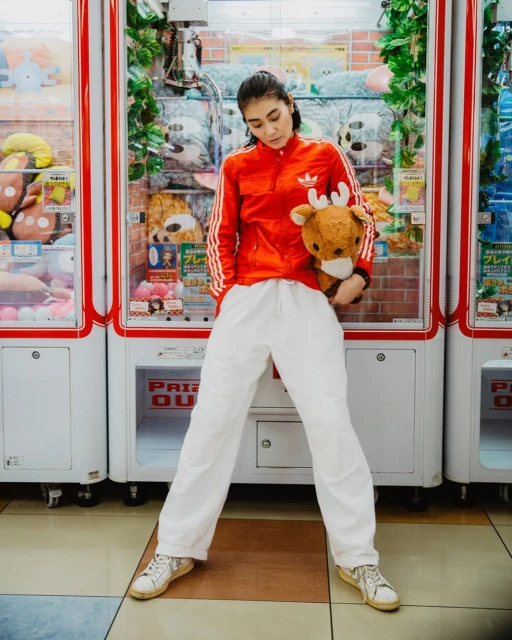 Young woman in red jacket poses with stuffed animal between arcade claw machines in an indoor gaming environment with bright lighting.
