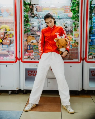 Young woman in red jacket poses with stuffed animal between arcade claw machines in an indoor gaming environment with bright lighting.