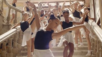 A group of diverse female ballet dancers in black leotards and white skirts practice on a grand staircase in natural light.