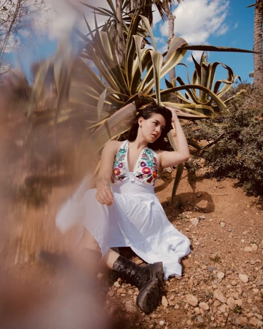 A woman in a white floral dress and black boots is lying in a natural setting with greenery, under a blue sky with light clouds.
